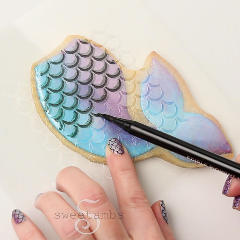 A finger is holding the mermaid scales template on the cookie while the other hand traces the stencil with an edible ink marker.