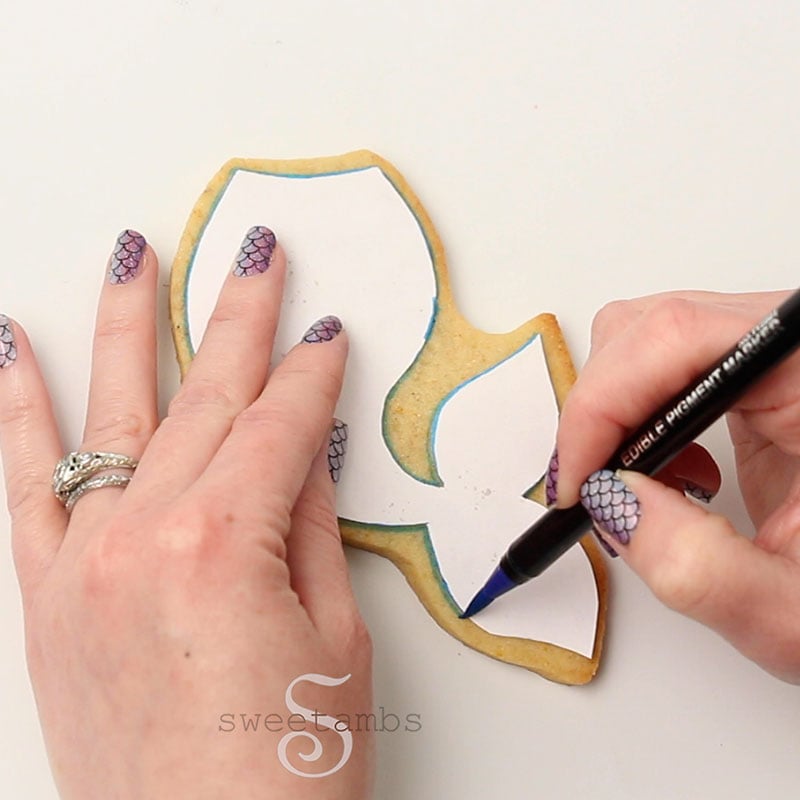 One hand is holding a paper mermaid tail template on a baked mermaid tail shaped cookie while the other hand traces the template with an edible ink marker