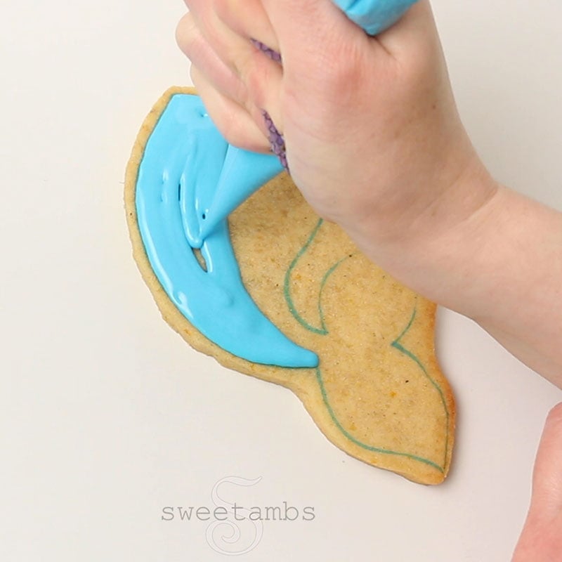 A hand is holding a piping bag filled with blue icing and  flooding half of the mermaid tail cookie.