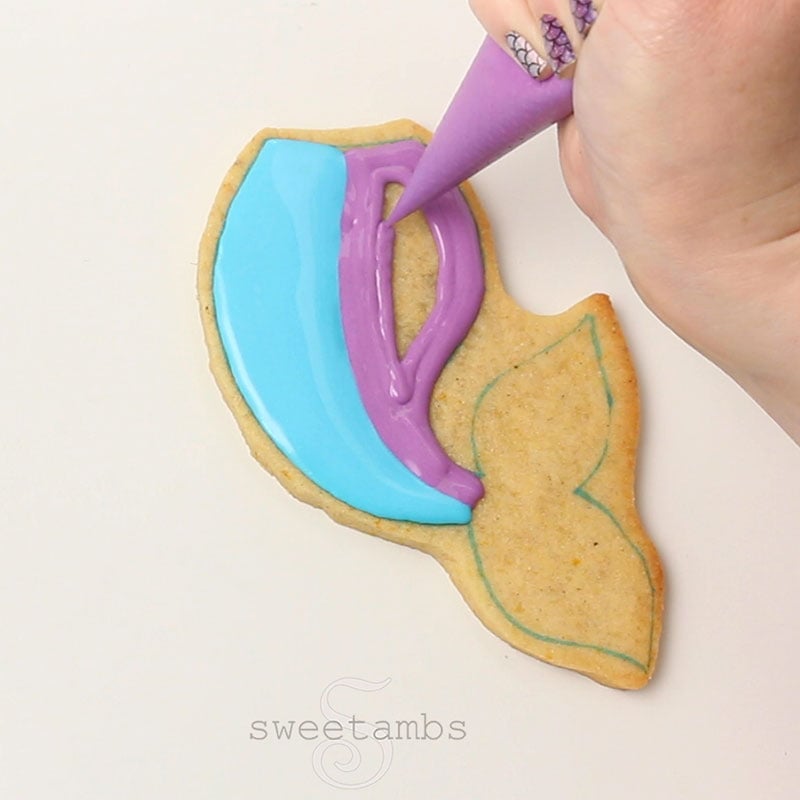 A hand is holding a piping bag filled with purple icing and flooding the other half of the mermaid tail cookie.