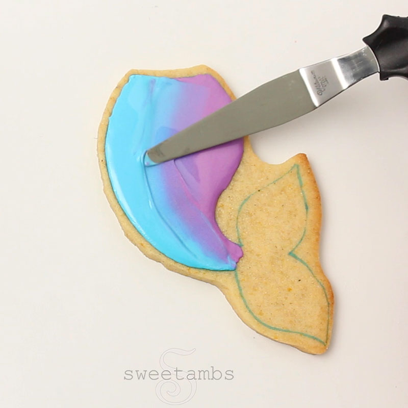 a tapered spatula is blending the blue and purple icing together in the middle of the mermaid tail cookie