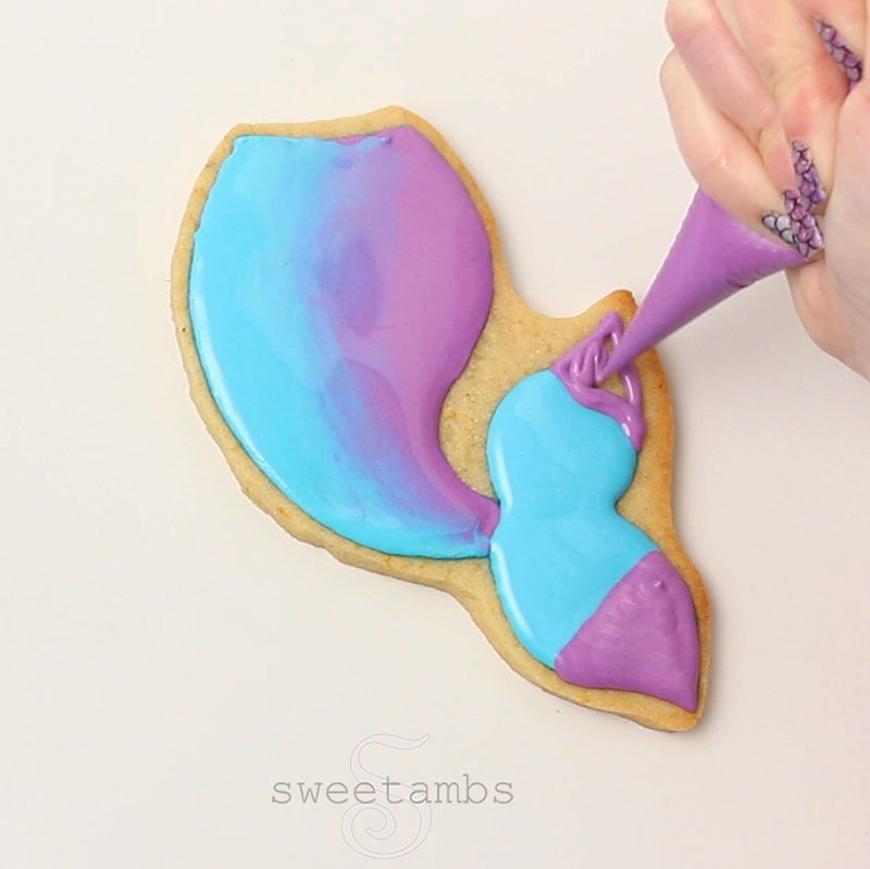 a hand is holding a piping bag filled with purple icing and is flooding the end of the fins of the mermaid tail cookie.