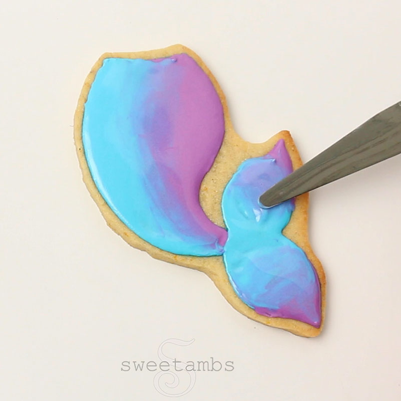 A tapered spatula is blending the blue and purple icing together on the fins of the mermaid tail cookie.