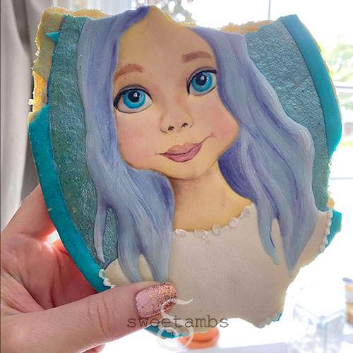 A portrait cookie of a child decorated to look like Elsa from Frozen 2. The cookie is being held up in front of a window and the cookie has bites taken out of it.