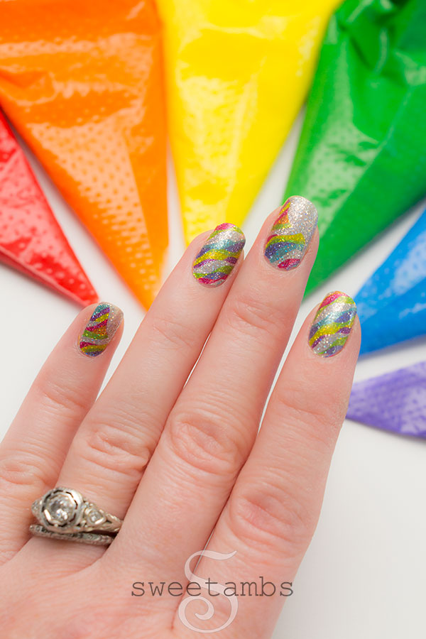 Sparkly rainbow nails over a background of rainbow icing in decorating bags. On the ring finger is an antique diamond ring.