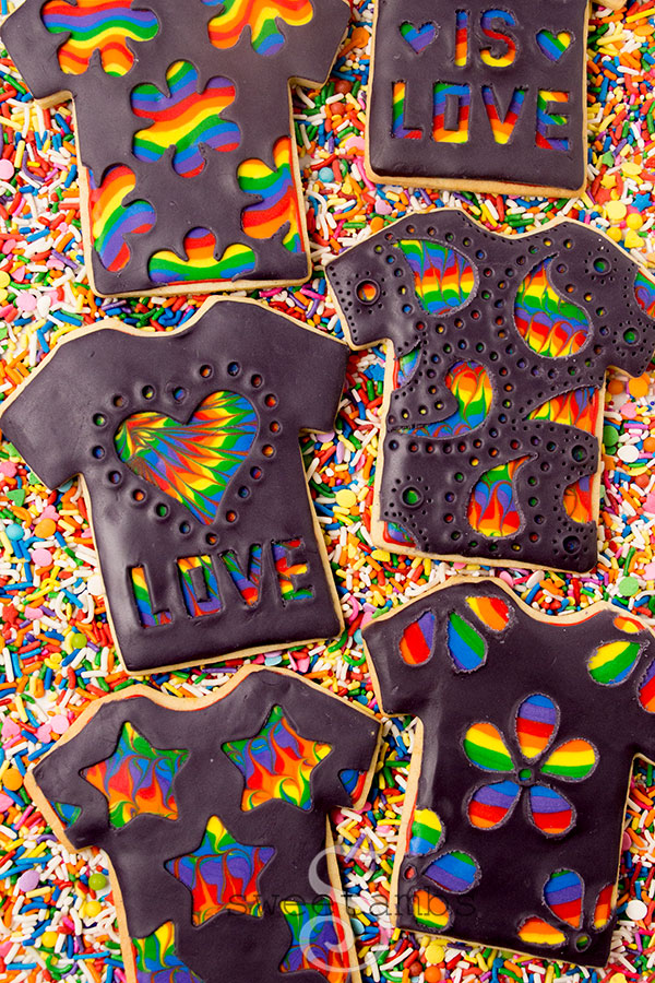 T-shirt cookies decorated with black modeling chocolate carved with designs that show rainbow patterns underneath. The cookies are laying on rainbow sprinkles.