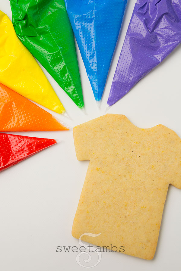 A rainbow of icing colors in tipless decorating bag surrounds a bare t-shirt shaped cookie.