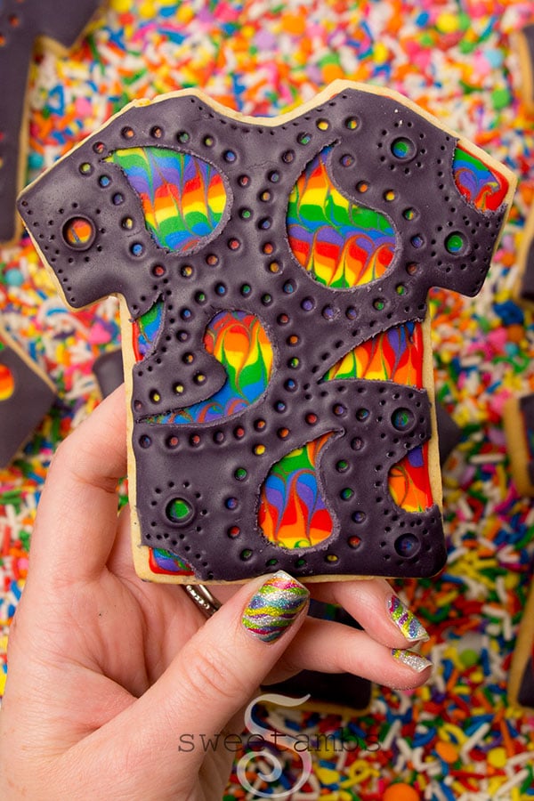 A t-shirt cookie covered in black modeling chocolate with a paisley design scratched into the modeling chocolate revealing a rainbow design underneath. A hand with sparkly rainbow nail polish is holding up the cookie. There is a background of rainbow sprinkles.