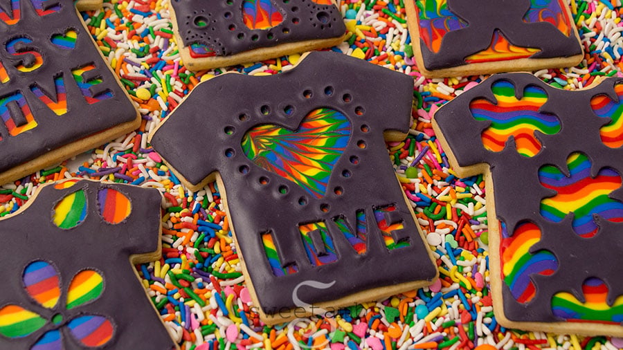 T-shirt cookies decorated with black modeling chocolate carved with designs that show rainbow patterns underneath. The cookies are laying on rainbow sprinkles.
