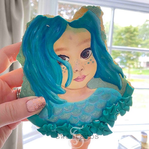 A portrait cookie of a small child decorated to look like a sea creature inspired by the movie Luca. The cookie is being held up in front of a window and the cookie has bites taken out of it.