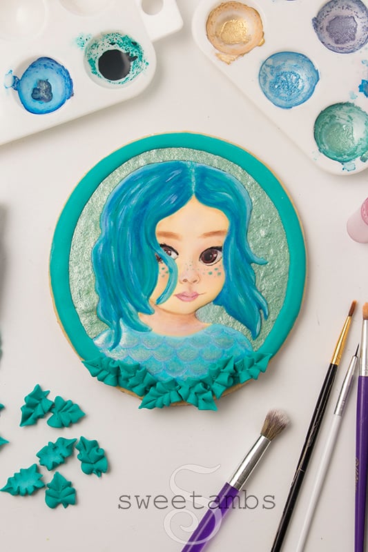 A portrait cookie of a small child decorated to look like a sea creature inspired by the movie Luca. The cookie is surrounded by painting supplies and royal icing leaves.