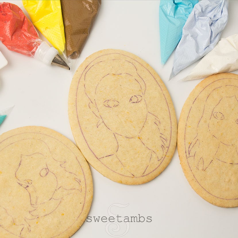 3 bare cookies with outlines of faces drawn on them with edible ink marker. There are piping bags filled with royal icing surrounding the cookies.