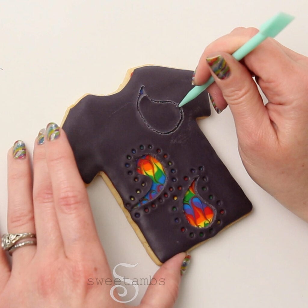 A hand is using the sharp end of a two-in-one tool to scratch a paisley design into the modeling chocolate. The other hand is holding the cookie in place.
