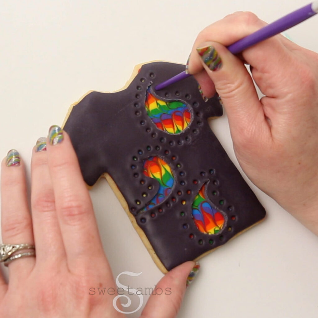 A hand is pushing the end of a handle of a brush into the modeling chocolate on the tshirt cookie to create a pattern of dots around the paisley. The other hand is holding the cookie in place.