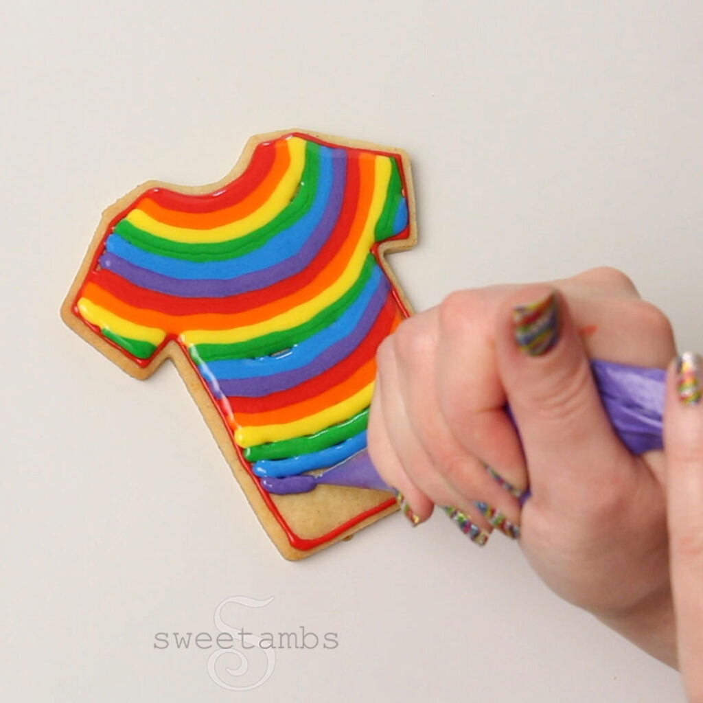 A hand with sparkly rainbow fingernails is piping a rainbow design in royal icing on the t-shirt cookie.