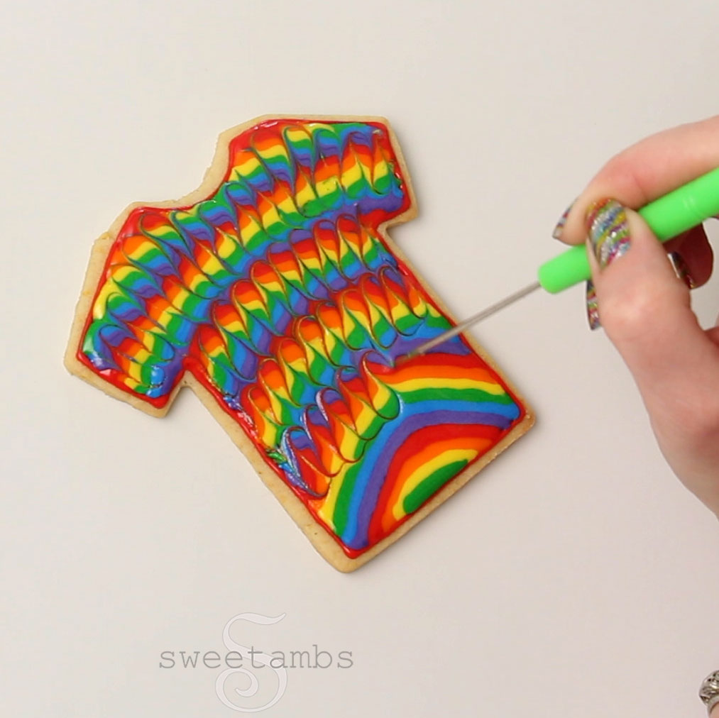 A hand with sparkly rainbow fingernails is dragging a scribe tool through the icing to create a wavy pattern in the rainbow royal icing.