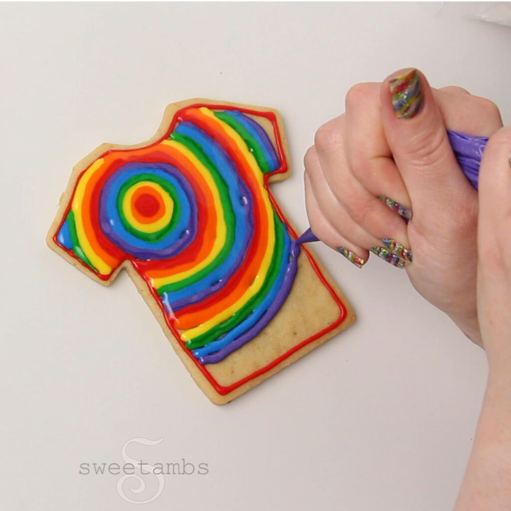 A hand with sparkly rainbow fingernails is piping a rainbow design in royal icing on the t-shirt cookie.