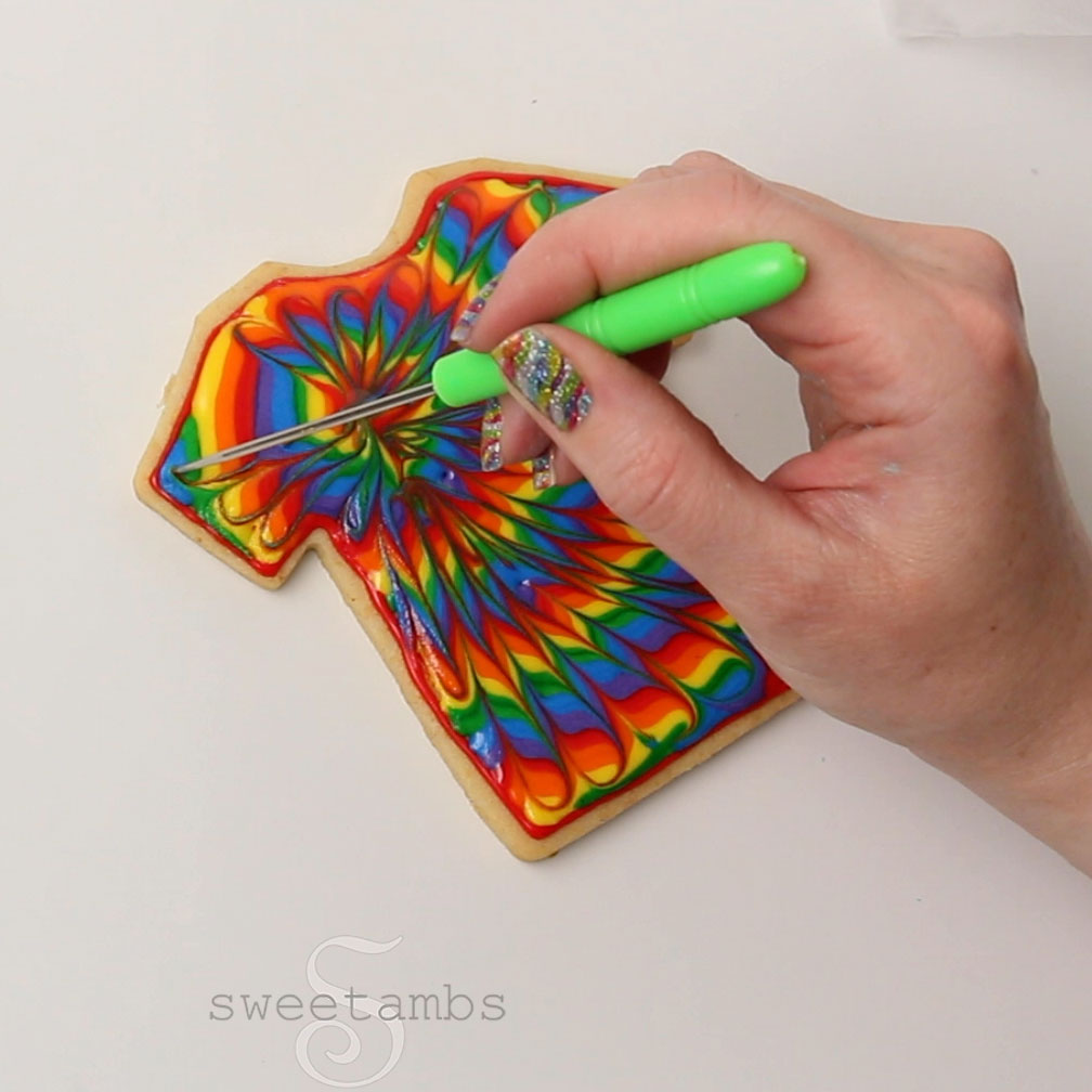 A hand with sparkly rainbow fingernails is dragging a scribe tool through the icing to create a tie dye effect in the rainbow royal icing.