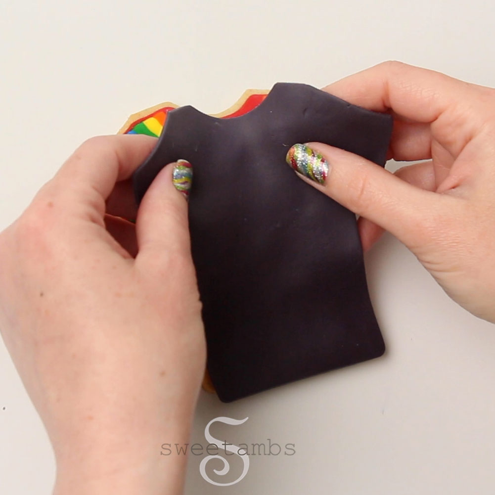 Two hands with sparkly rainbow nails are placing the modeling chocolate t-shirt over the t-shirt cookie with a rainbow pattern.