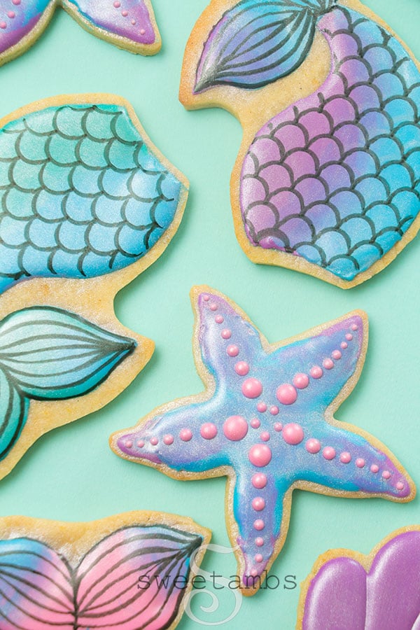 a set of cookies decorated to look like mermaid tails and starfish. They are iced with teal, purple, blue, and pink icing with a coating of shimmery luster dust. They are on a teal background. 