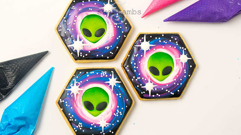 3 hexagon shaped cookies decorated with royal icing aliens and a galaxy design. There are 4 piping bags around the cookies filled with black, blue, pink, and purple royal icing.
