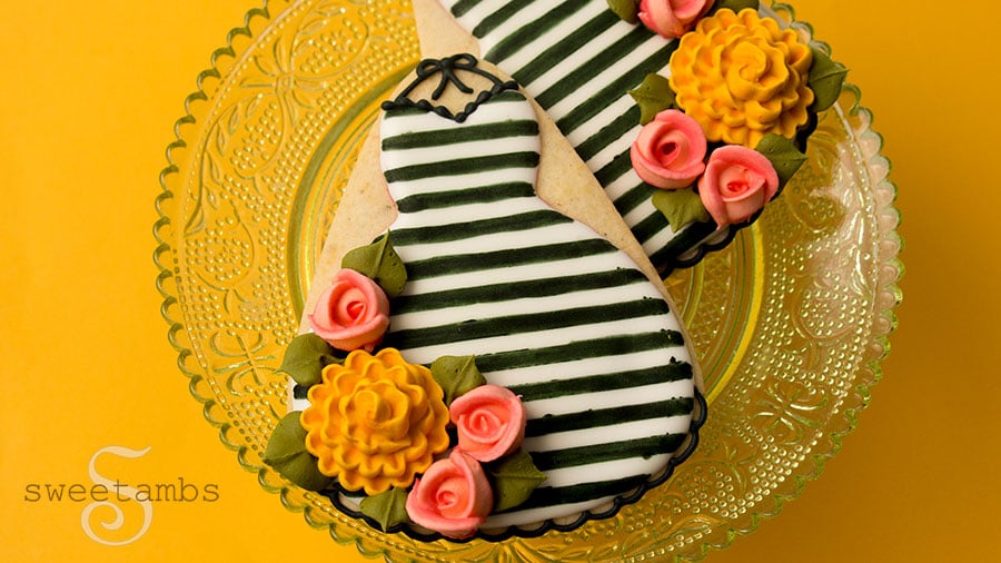 A set of cookies decorated to look like fall dresses. The cookies are decorated in black and white stripes with pink royal icing roses and golden yellow royal icing chrysanthemums. They are on a transparent light green cake plate with a lace pattern over a golden yellow background.