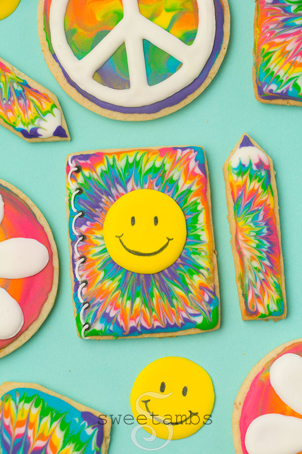 Notebook and pencil shaped cookies decorated with groovy royal icing tie dye designs. The notebook has a yellow smiley face. The notebook and pencil are surrounded by other cookies including a peace sign cookie.