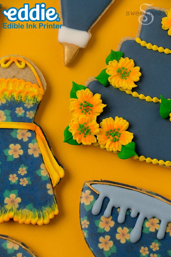 a set of cookies decorated to look like a tiered cake, a piping bag, tiny cookies, an apron, and a stand mixer. The cookies are decorated with golden yellow flowers and dark blue icing. They are on a golden yellow background.