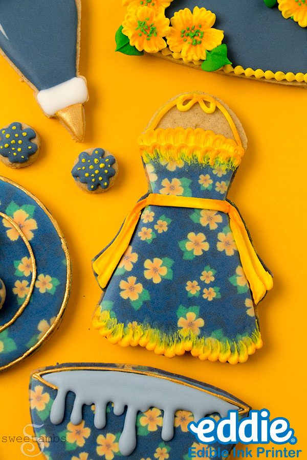 A set of decorated cookies that look like a piping bag icing small flowers, an apron, and a bowl filled with blue icing. The cookies are decorated with dark blue icing and golden yellow flowers. They are on a golden yellow background.