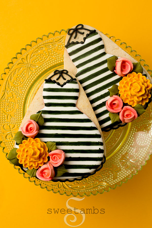 Een set koekjes versierd om eruit te zien als herfstjurken.  De koekjes zijn gedecoreerd in zwart-witte strepen met roze royal icing rozen en goudgele royal icing chrysanten.  Ze staan ​​op een transparant lichtgroen taartbord met een kantpatroon op een goudgele achtergrond.