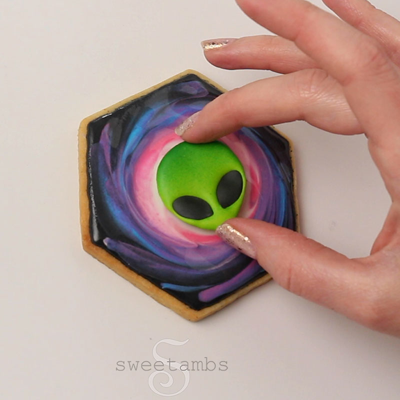 a hand is placing the alien face royal icing transfer onto the freshly iced cookie