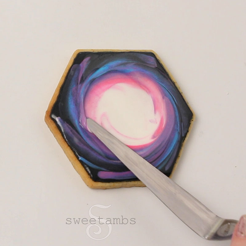 a tapered spatula is blending the royal icing colors in a swirl motion on the hexagon cookie.