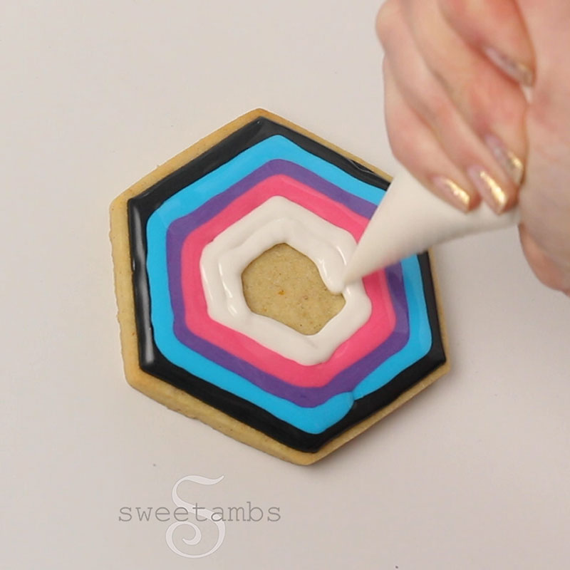 A piping bag filled with white royal icing is icing a hexagon shaped cookie. The cookie is already iced with rings of black, blue, purple, and pink royal icing.