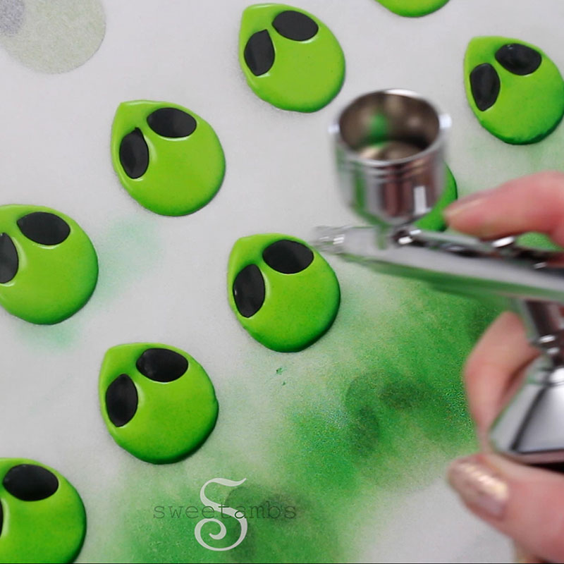 An airbrush is adding darker green shading to the edges of the royal icing alien faces.