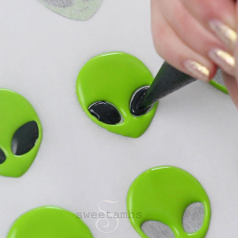 A piping bag filled with black royal icing is filling in the eyes on the alien's face.