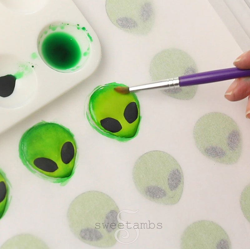 A paintbrush is applying green food coloring to add shading to the royal icing alien faces.