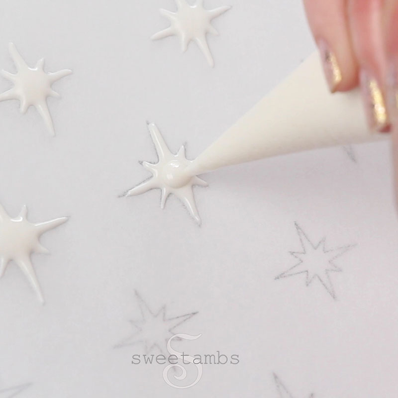 a piping bag filled with white royal icing is filling in stars on a piece of parchment paper over a star template.