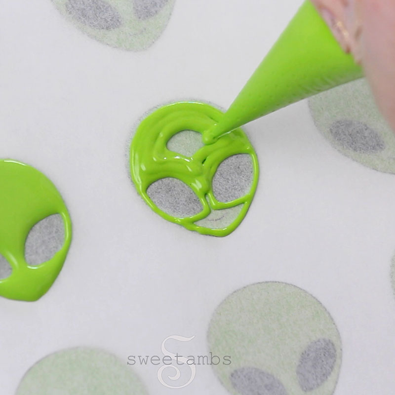 A piping bag filled with bright green royal icing is filling in an alien face following a template. 