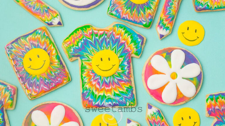 A set of cookies decorated with a rainbow tie dye icing design. The cookies are shaped like t-shirts, notebooks, and pencils, and decorated with smiley faces and flowers.