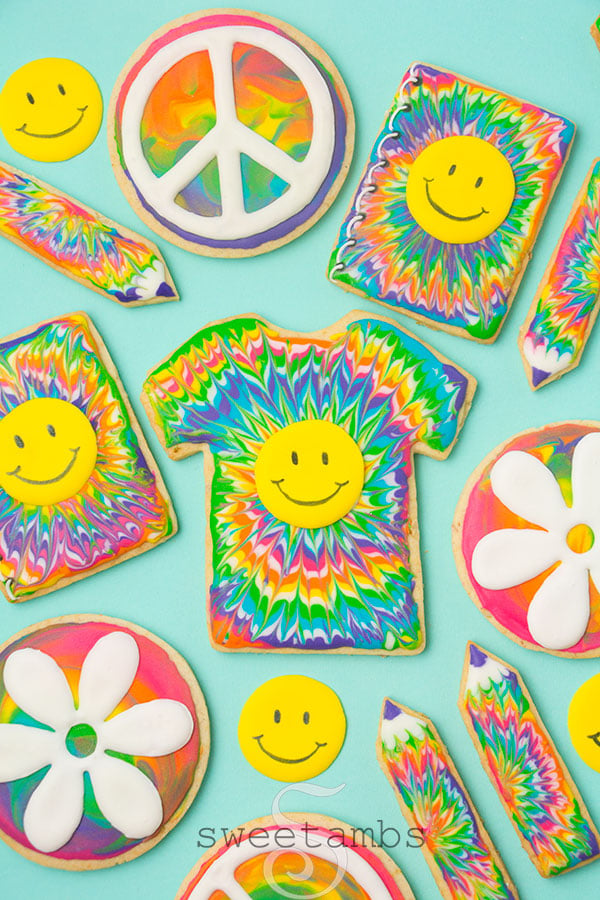 A set of cookies decorated with a rainbow tie dye icing design. The cookies are shaped like t-shirts, notebooks, and pencils, and decorated with smiley faces, peace signs, and flowers.