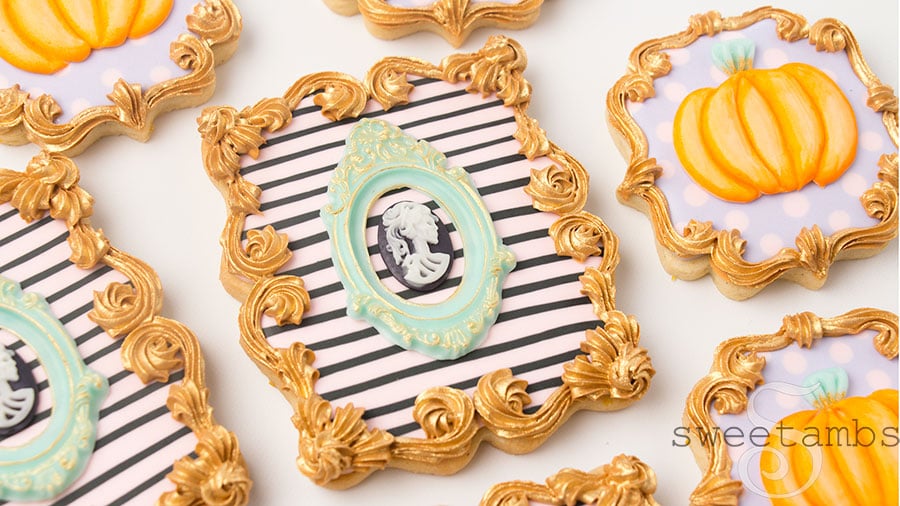 A set of plaque shaped cookies decorated with glam halloween designs. The larger plaque cookie is decorated with light pink icing with black stripes, a teal green frame and a black and white skull cameo in the center. There is a gold ornate border on the cookies. the smaller plaque cookie is decorated with light purple icing and light pink polka dots with an orange pumpkin and a teal stem.