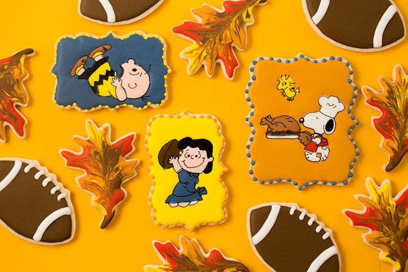 Decorated cookies depicting a scene from A Charlie Brown Thanksgiving where Lucy pulls a football out from under Charlie Brown. Snoopy is carrying a turkey and Woodstock is flying nearby. There are also marbled fall leaves and football cookies.