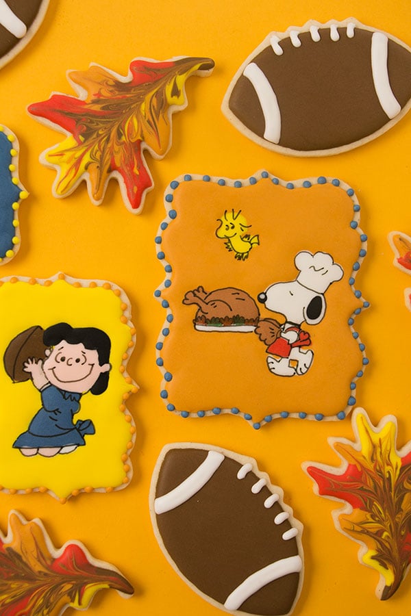 Decorated cookies depicting a scene from A Charlie Brown Thanksgiving where Lucy pulls a football out from under Charlie Brown. Snoopy is carrying a turkey and Woodstock is flying nearby. There are also marbled fall leaves and football cookies.