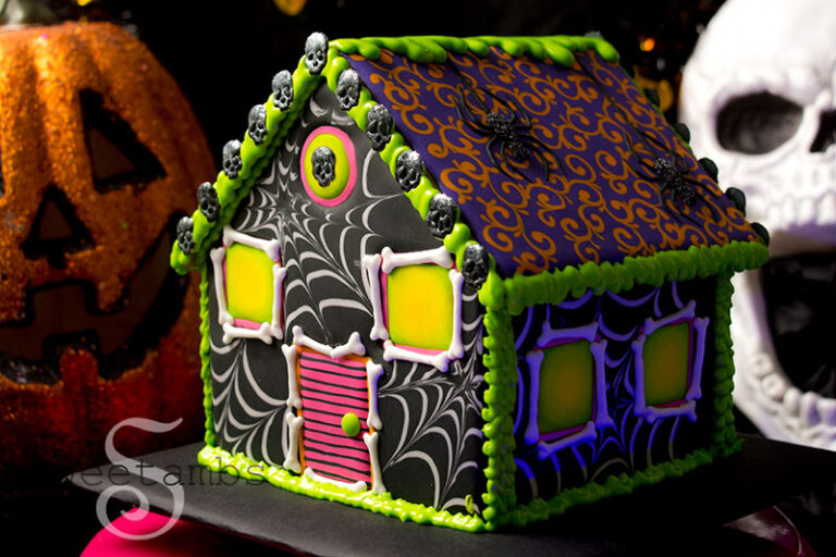 A halloween gingerbread house decorated with black and neon colored royal icing. The front and sides are decorated with a spiderweb design and the roof is decorated with a filigree design. There is a jack o lantern and a skull behind the house.