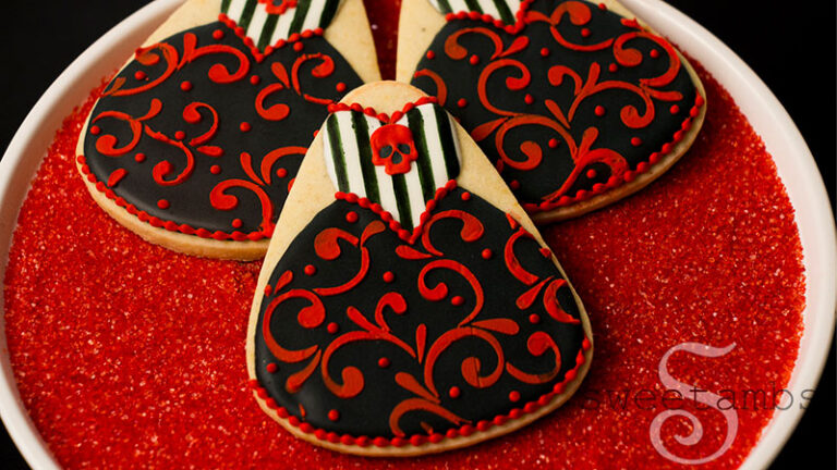 Red and black Halloween gown cookies decorated with filigree, stripes, and royal icing skulls. The cookies are sitting on a platter filled with red sanding sugar.