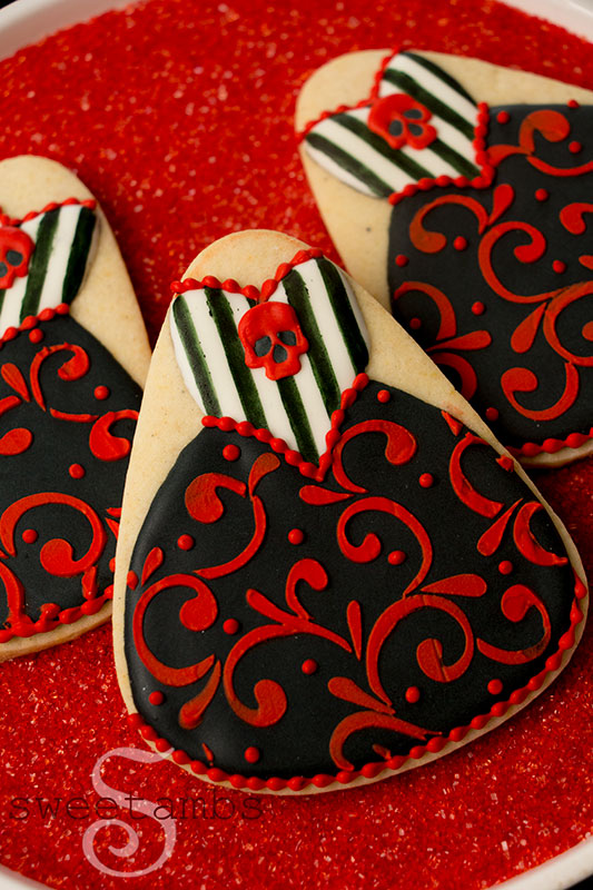 Red and black Halloween gown cookies decorated with filigree, stripes, and royal icing skulls. The cookies are sitting on a platter filled with red sanding sugar.