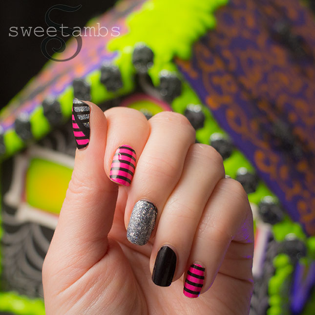 Fingernails decorated with pink and black stripes, silver glitter, and black nail polish. The Halloween gingerbread house is in the background.