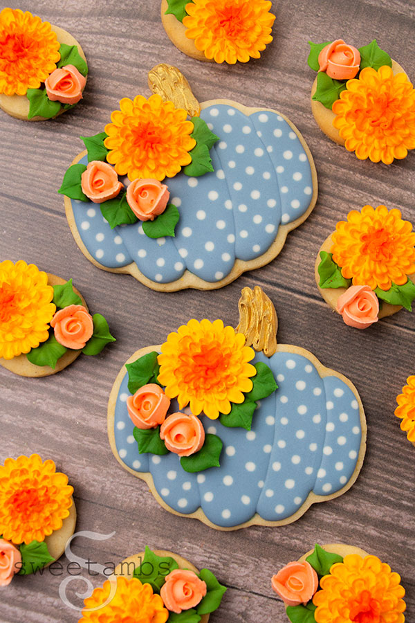 pumpkin shaped cookies decorated with light blue royal icing white white polka dots. The pumpkin cookies have gold stems and are also decorated with royal icing roses and chrysanthemums. The cookies are surrounded by more royal icing flowers and are on a wooden background.