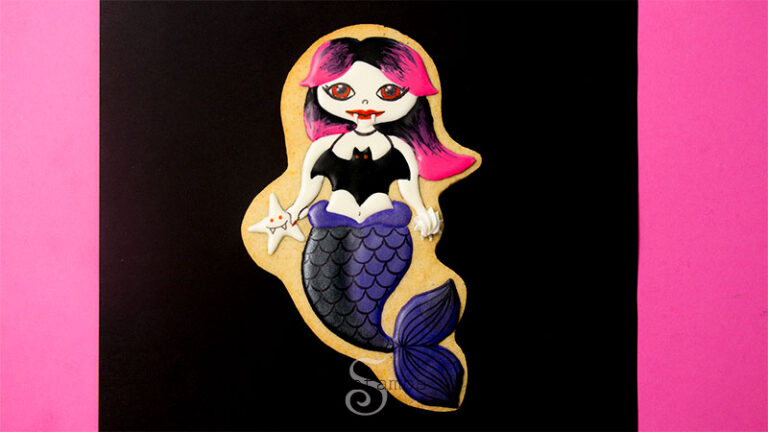 A cookie decorated to look like a vampire mermaid with a black and purple tail and pink and black hair. The cookie is on a black and hot pink background