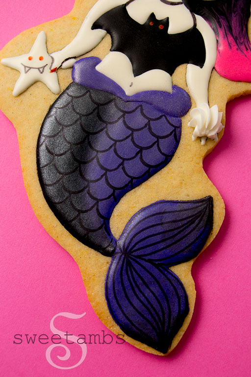 Een close-up van een koekje dat is versierd om eruit te zien als een vampierzeemeermin met een zwarte en paarse staart en roze en zwart haar.  Het koekje is op een hete roze achtergrond.  De vampiermeermin houdt een vampierzeester in de ene hand en een zeeschelp in de andere.